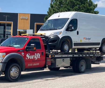 A red Swift Recovery tow truck transporting a white Budget rental van on its flatbed in front of a Jeep dealership.