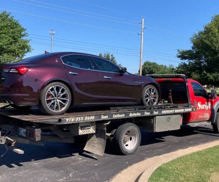 A maroon Maserati sedan is loaded on the back of a red flatbed tow truck parked on a street.