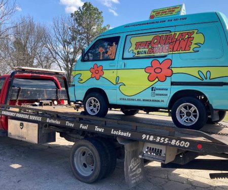 A red tow truck carries a van decorated like the Mystery Machine from Scooby-Doo on its flatbed.