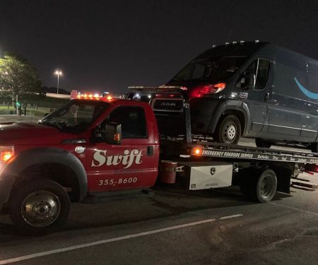 A red tow truck is transporting a dark blue Amazon Prime delivery van at night in a parking lot.